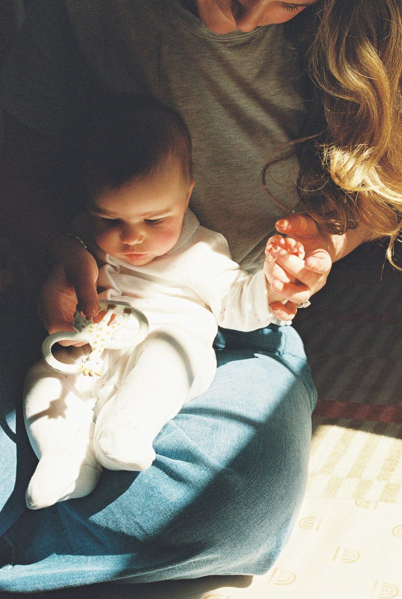 A mother holds her infant baby in her lap, bathed in warm sunlight, as the child plays with a white shoe.