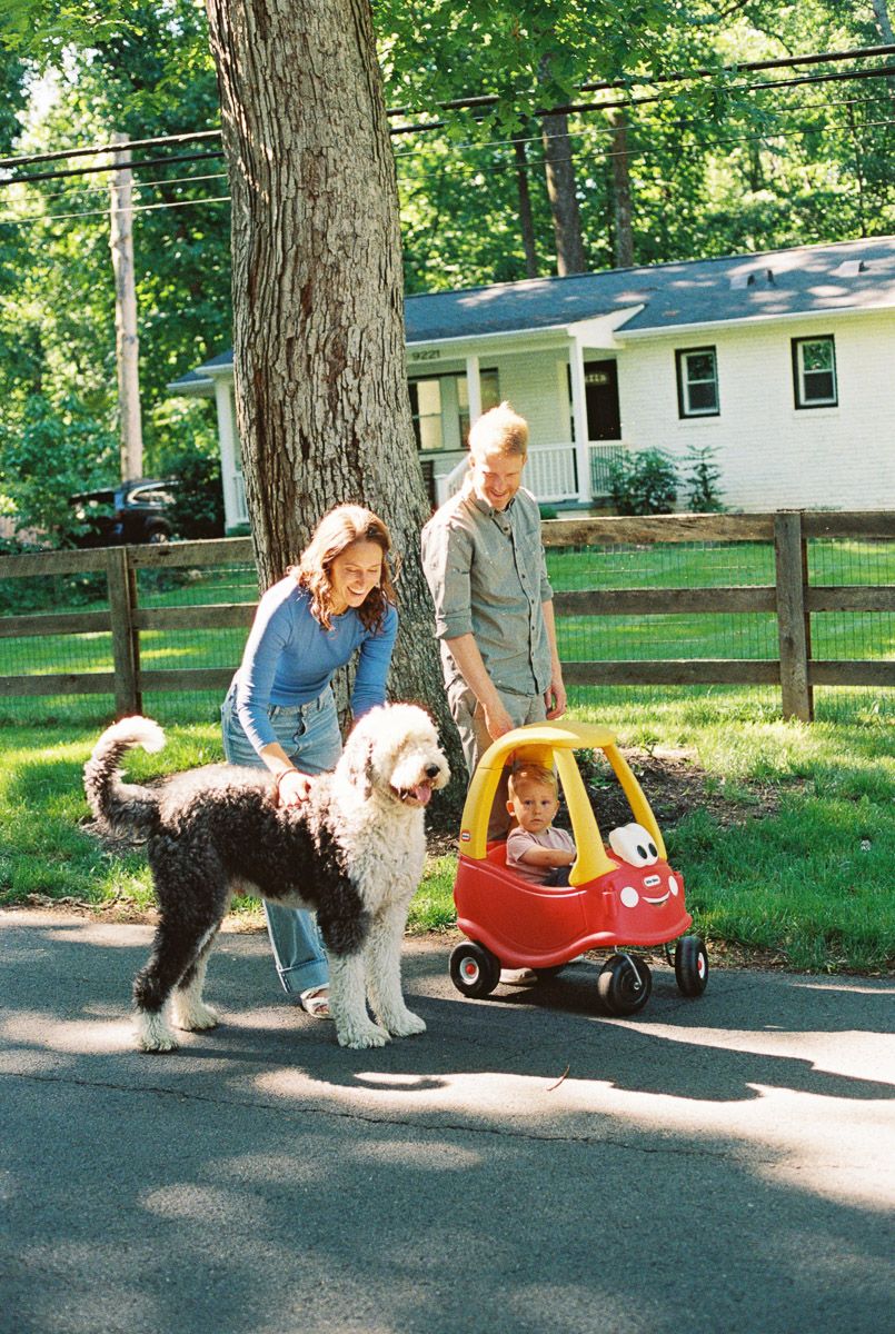 Family with large shaggy dog and toddler in red Little Tikes Cozy Coupe toy car on suburban driveway.