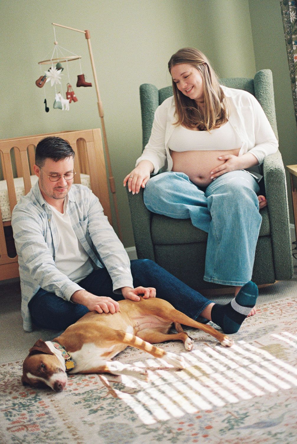 Expecting couple smiling with dog in nursery, woman seated in glider chair touching baby bump.