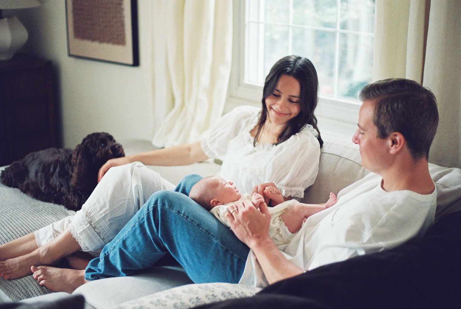 A young family relaxes on a couch, with a woman and man lovingly holding their newborn baby near a bright window.