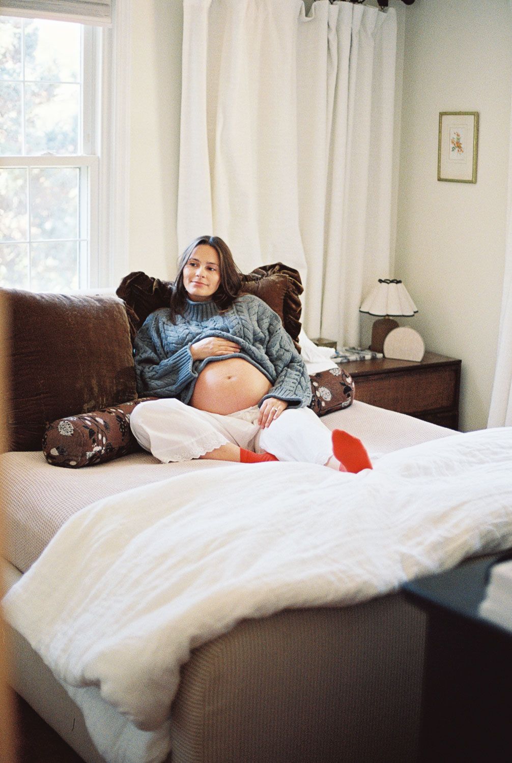 Pregnant woman sitting on a bed in a cozy bedroom, wearing a blue sweater with her baby bump exposed.