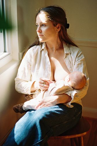 A mother breastfeeding her newborn baby while seated by a sunlit window, wearing a white shirt and jeans.