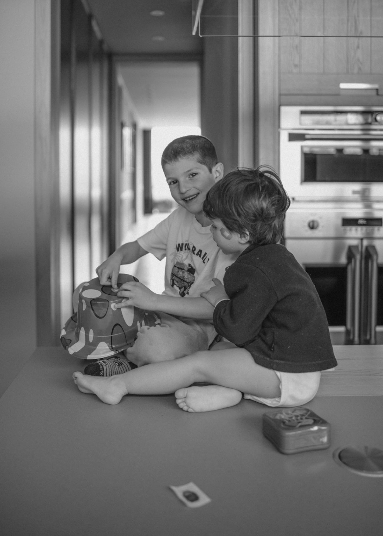 Two children sit together on a kitchen counter sharing a playful moment in black and white photography.