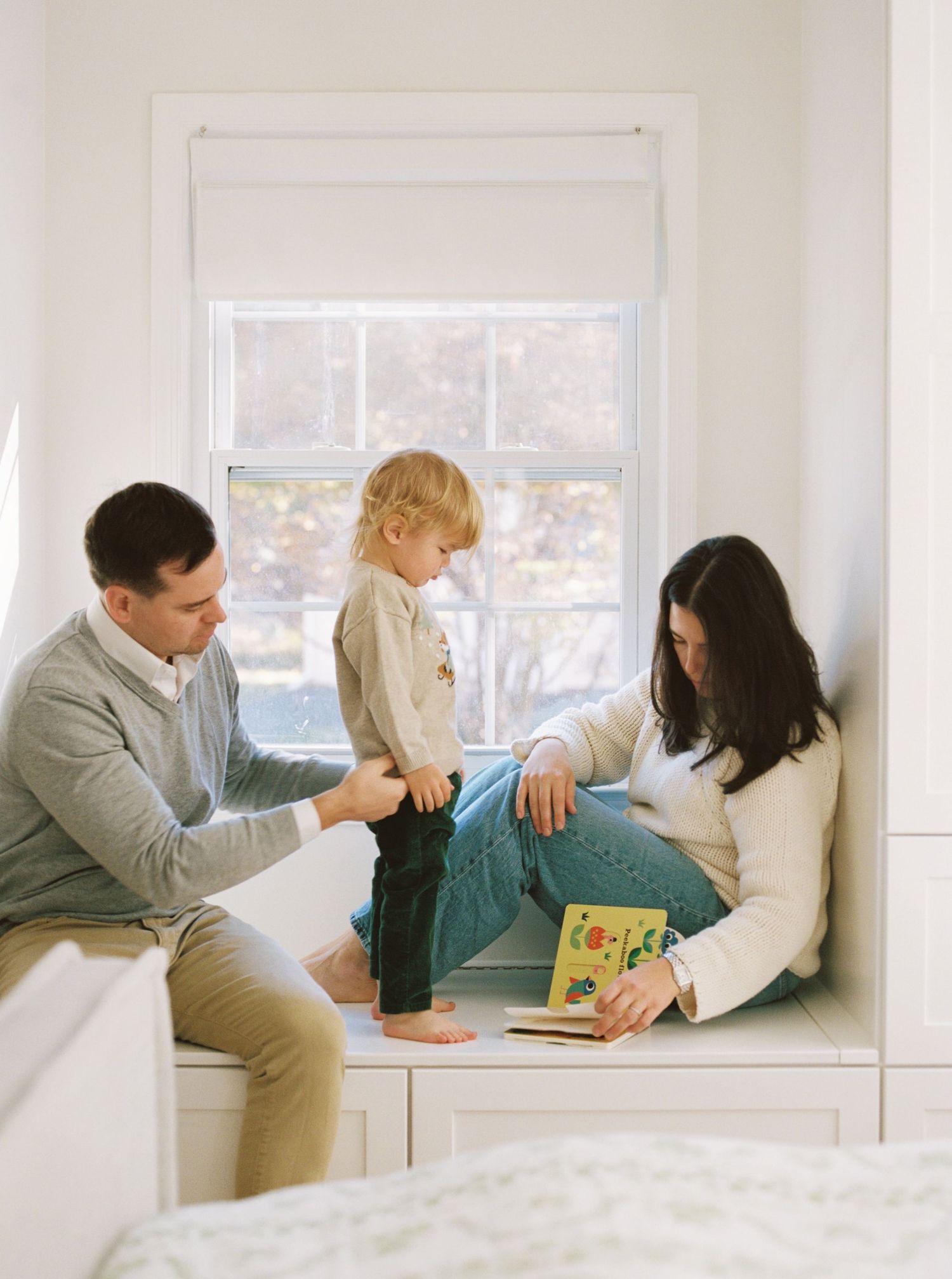 A family sits together in a sunny window nook reading a children's book.
