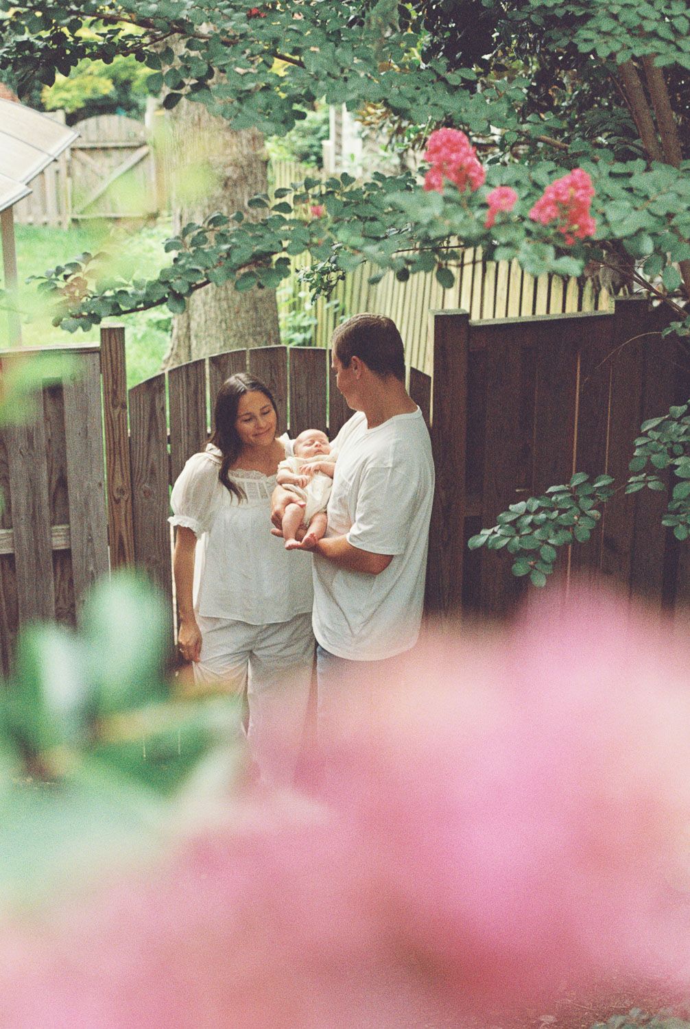 A couple stands in a lush garden holding their newborn baby, surrounded by pink flowers and a wooden fence.
