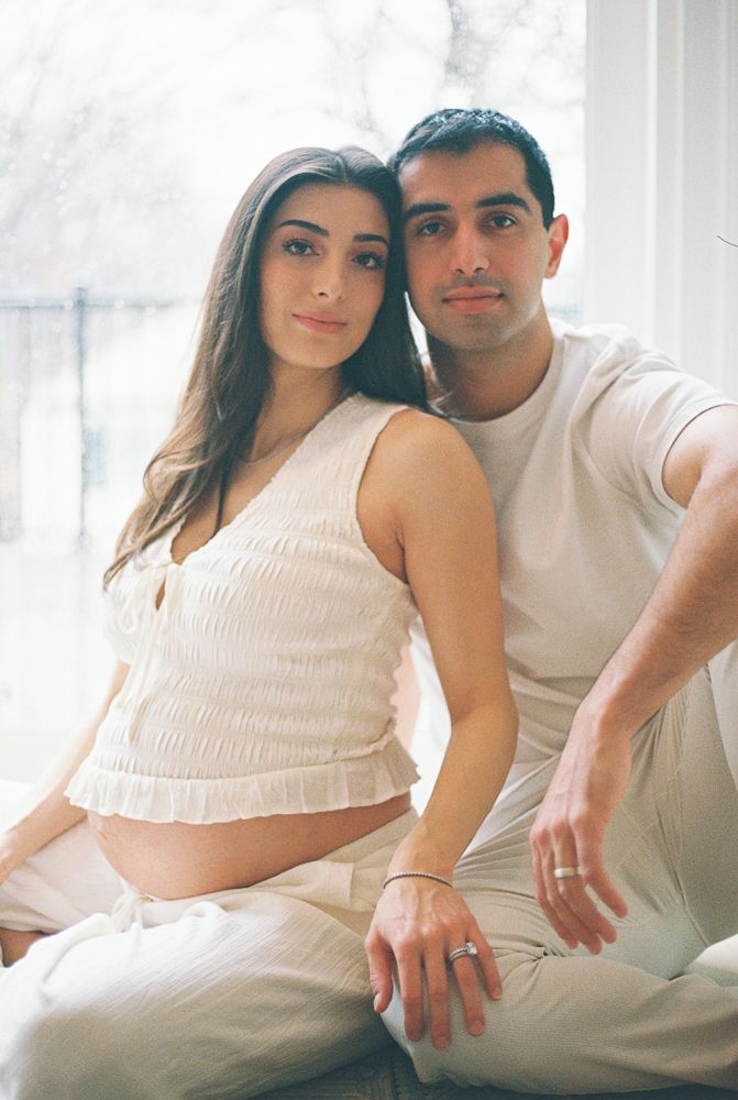 A couple in white outfits poses together on a couch in a bright room for a maternity photoshoot.