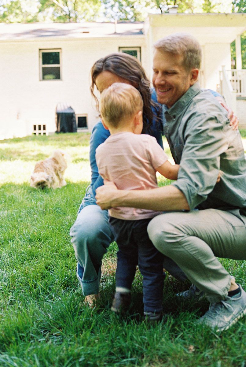 A smiling family with a toddler crouching on green grass in a backyard with a fluffy dog nearby.