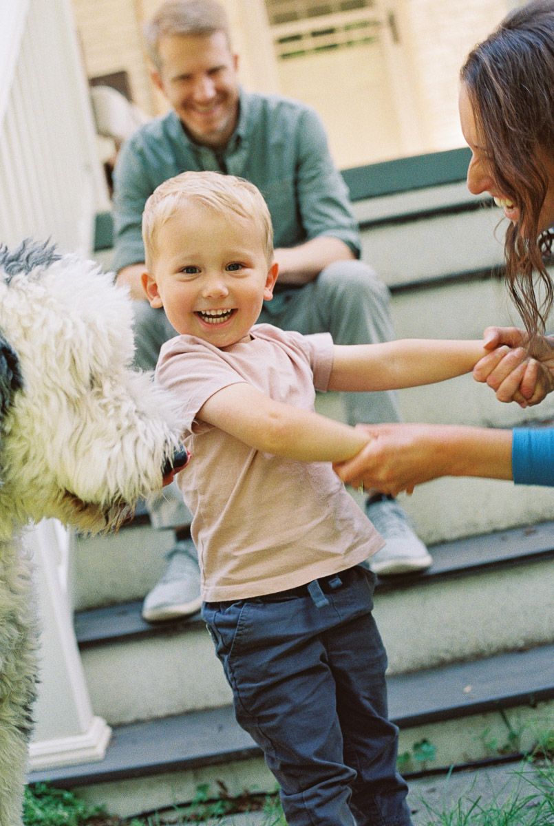 Happy toddler boy smiling on porch steps with parents and a fluffy white dog in a family portrait.