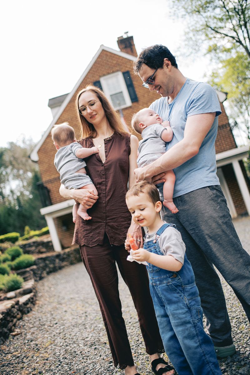 A smiling family with two babies and a toddler in overalls stands outside their brick home on a gravel path.