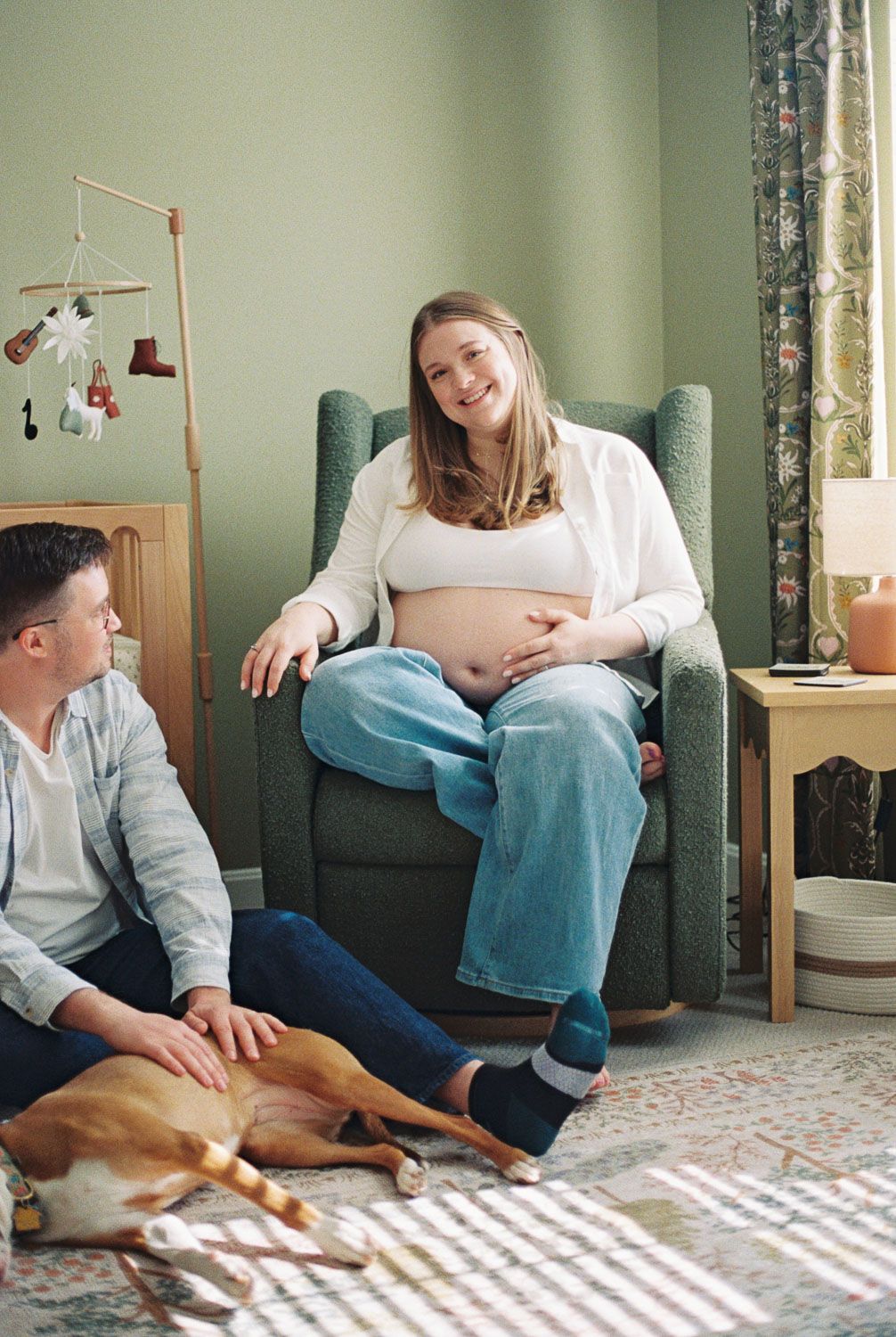 Smiling pregnant woman in armchair touching belly, with partner and dog in cozy nursery room.