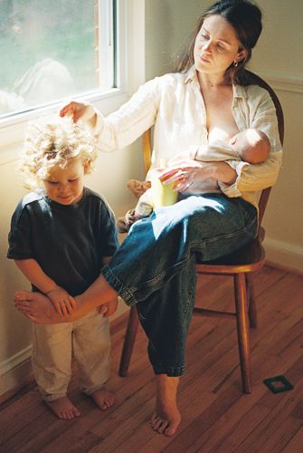 Mother breastfeeding newborn while seated on wooden chair, gently touching toddler's curly hair by sunny window.