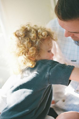 A curly-haired toddler in a teal shirt interacts closely with a man in a blue plaid shirt in soft natural light.
