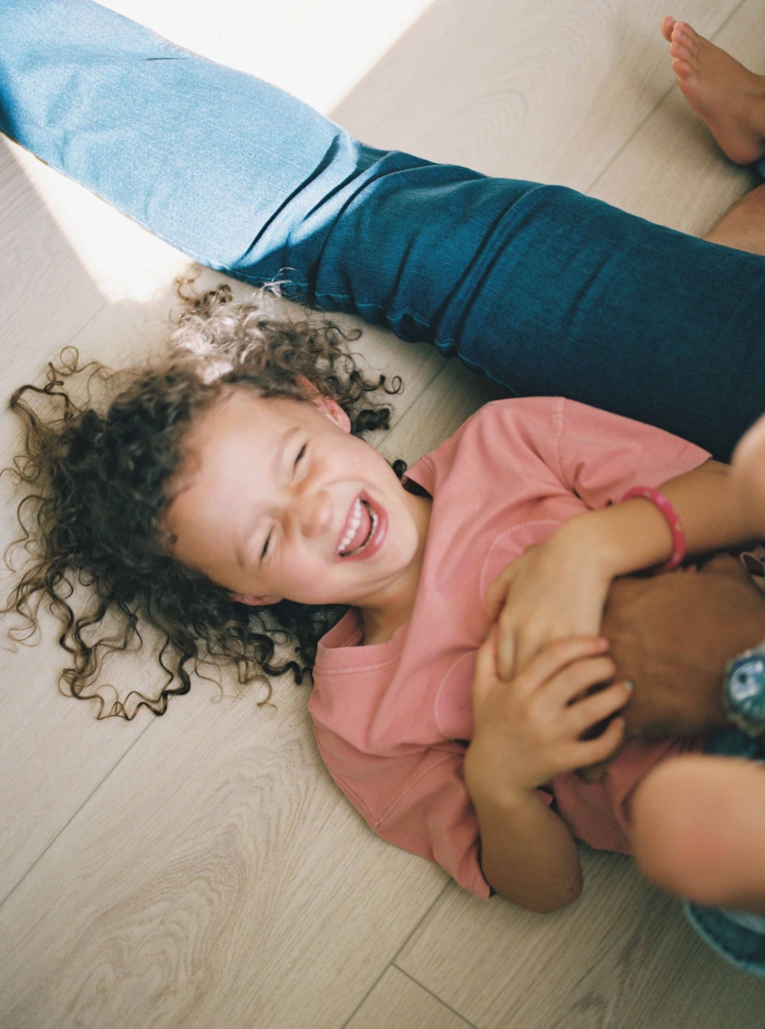 Person with curly hair lying on floor wearing pink sweater and laughing joyfully.