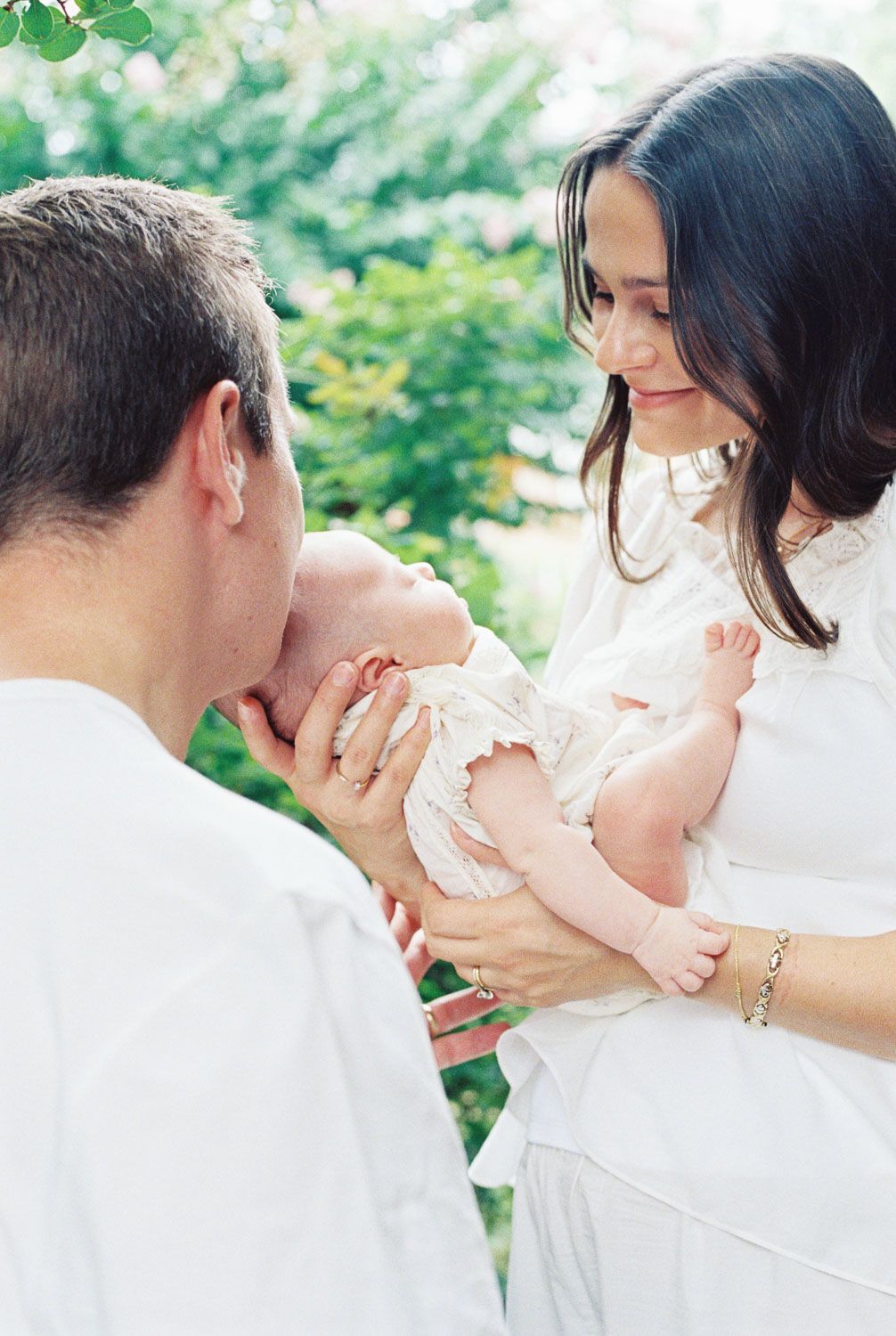 A couple in white clothing tenderly gazes at their newborn baby outdoors surrounded by lush green foliage.