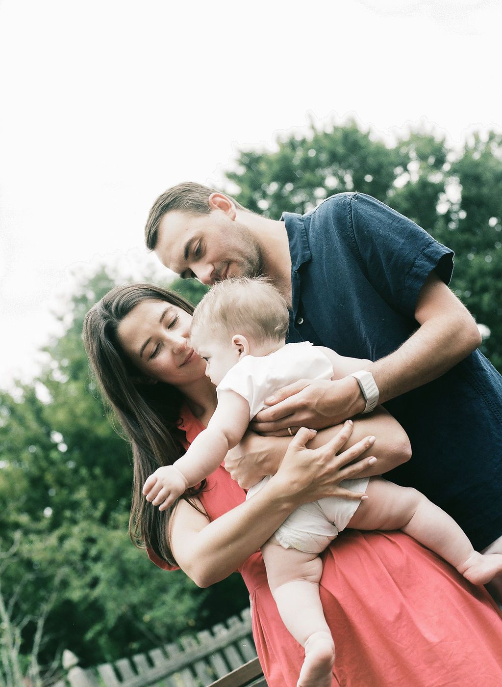 A loving family embraces their small baby outdoors on a sunny day with green trees in the background.