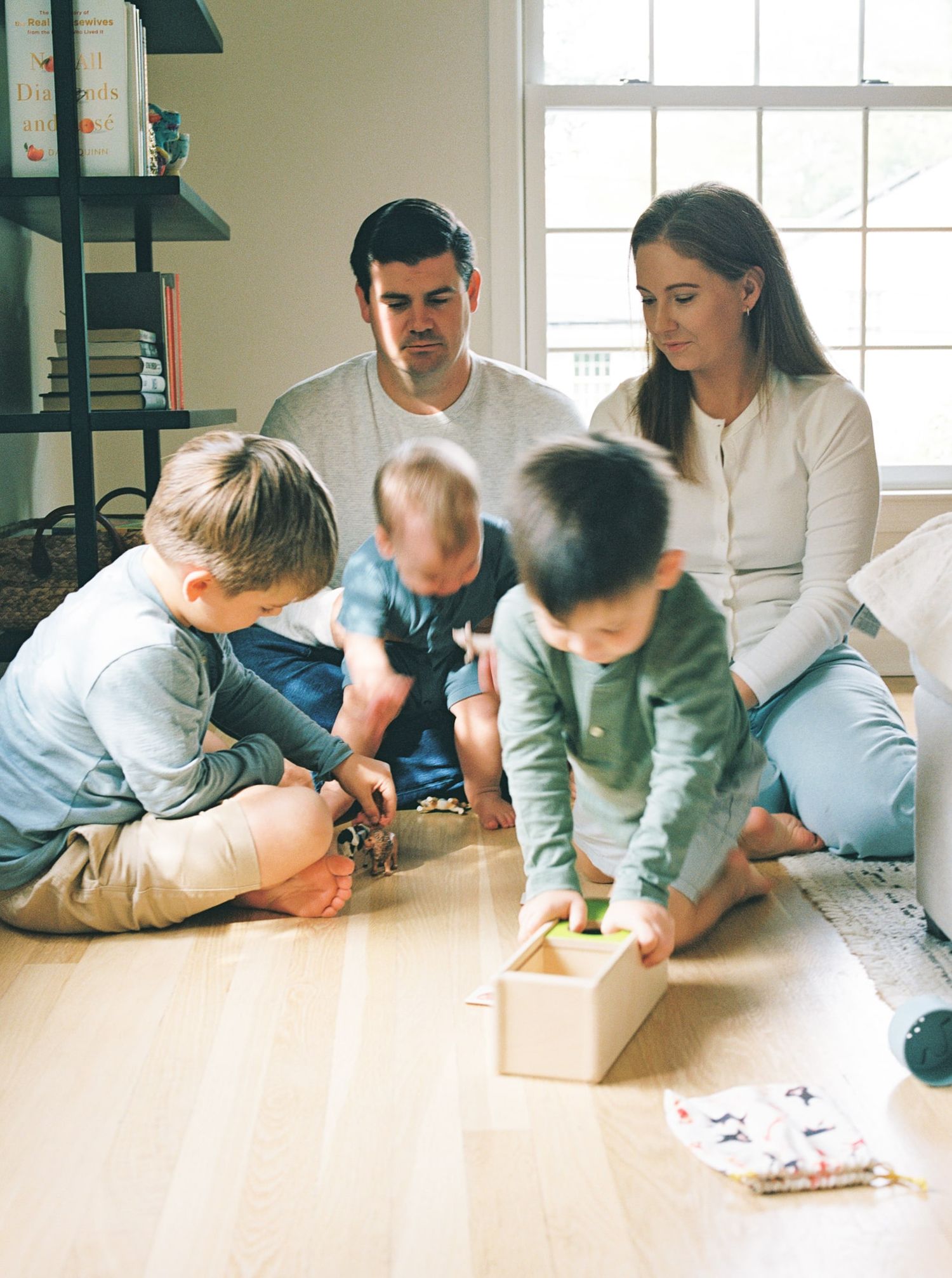 A group sits together on a hardwood floor playing with a wooden toy box in a bright room with bookshelves.