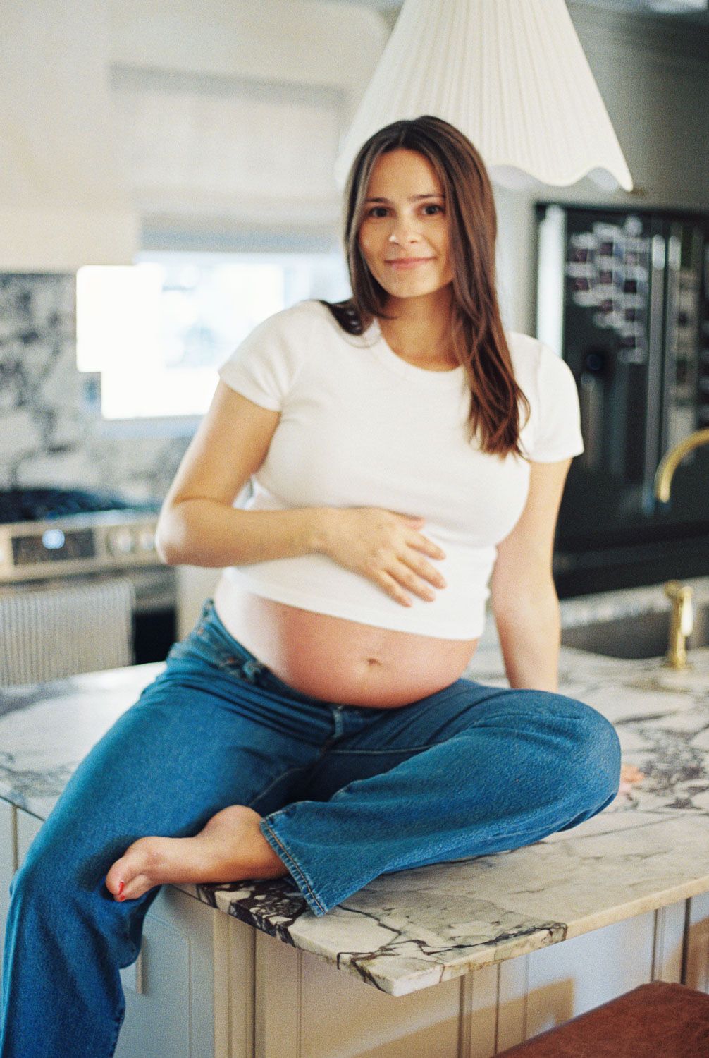 Pregnant woman in white crop top and jeans sitting cross-legged on kitchen counter, smiling and cradling her baby bump.
