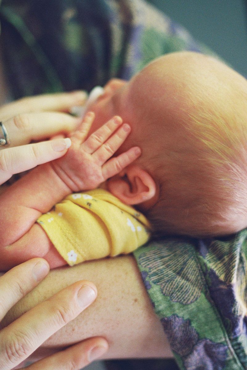 Close-up of a newborn baby with a tiny hand resting on its cheek, wrapped in a yellow blanket and held in a parent's arms.