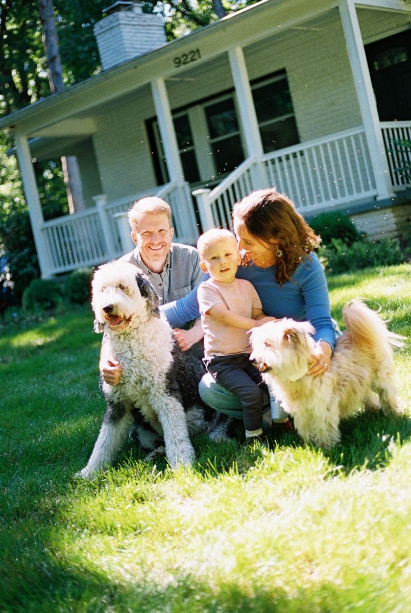 Family with toddler and two fluffy dogs smiling on green lawn in front of white house with porch.