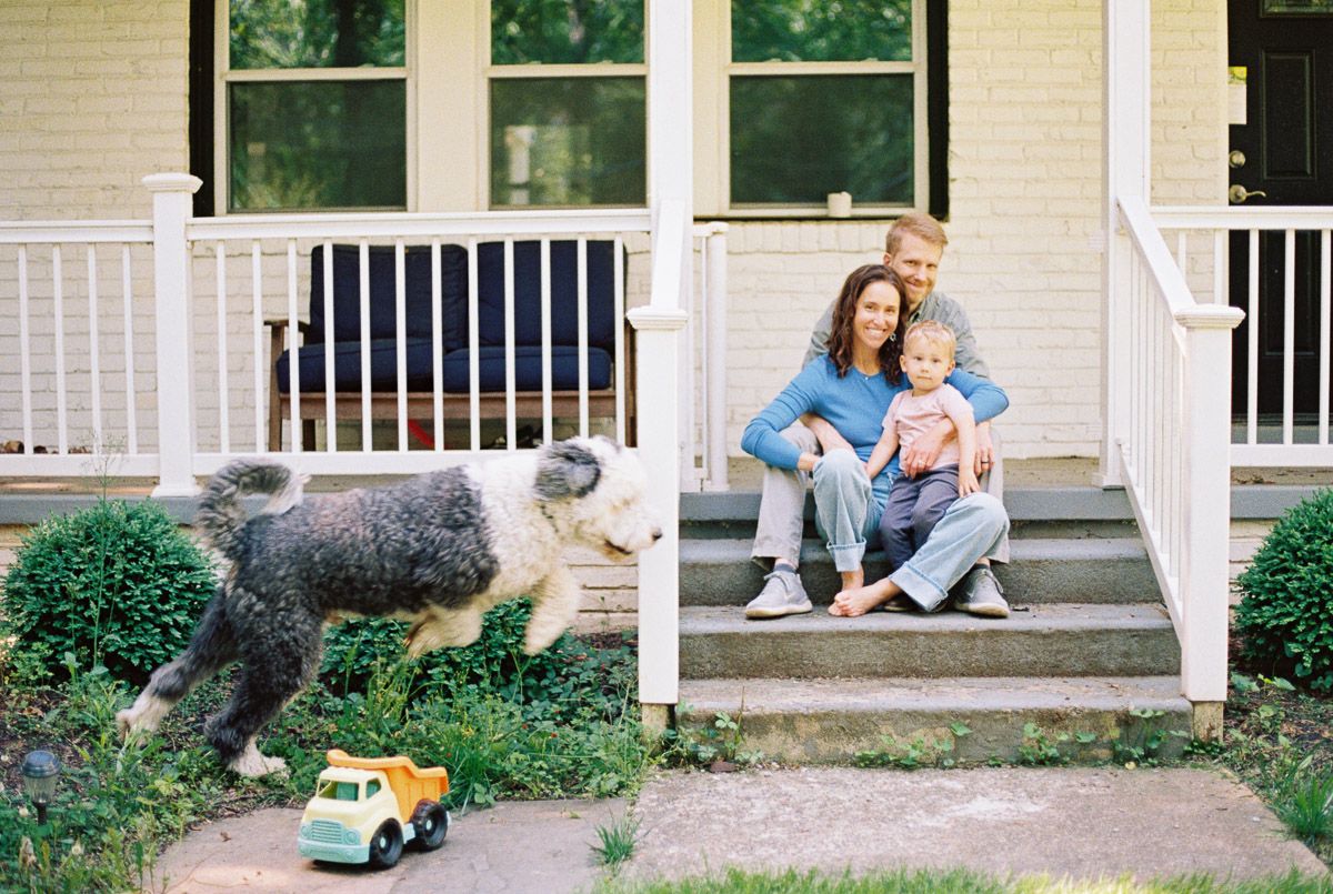 Family of three sitting on porch steps with fluffy dog and yellow toy truck in front of white house.