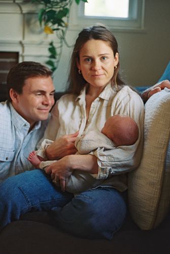Young couple sitting on a couch together, smiling, while the woman holds a newborn baby in their cozy home.