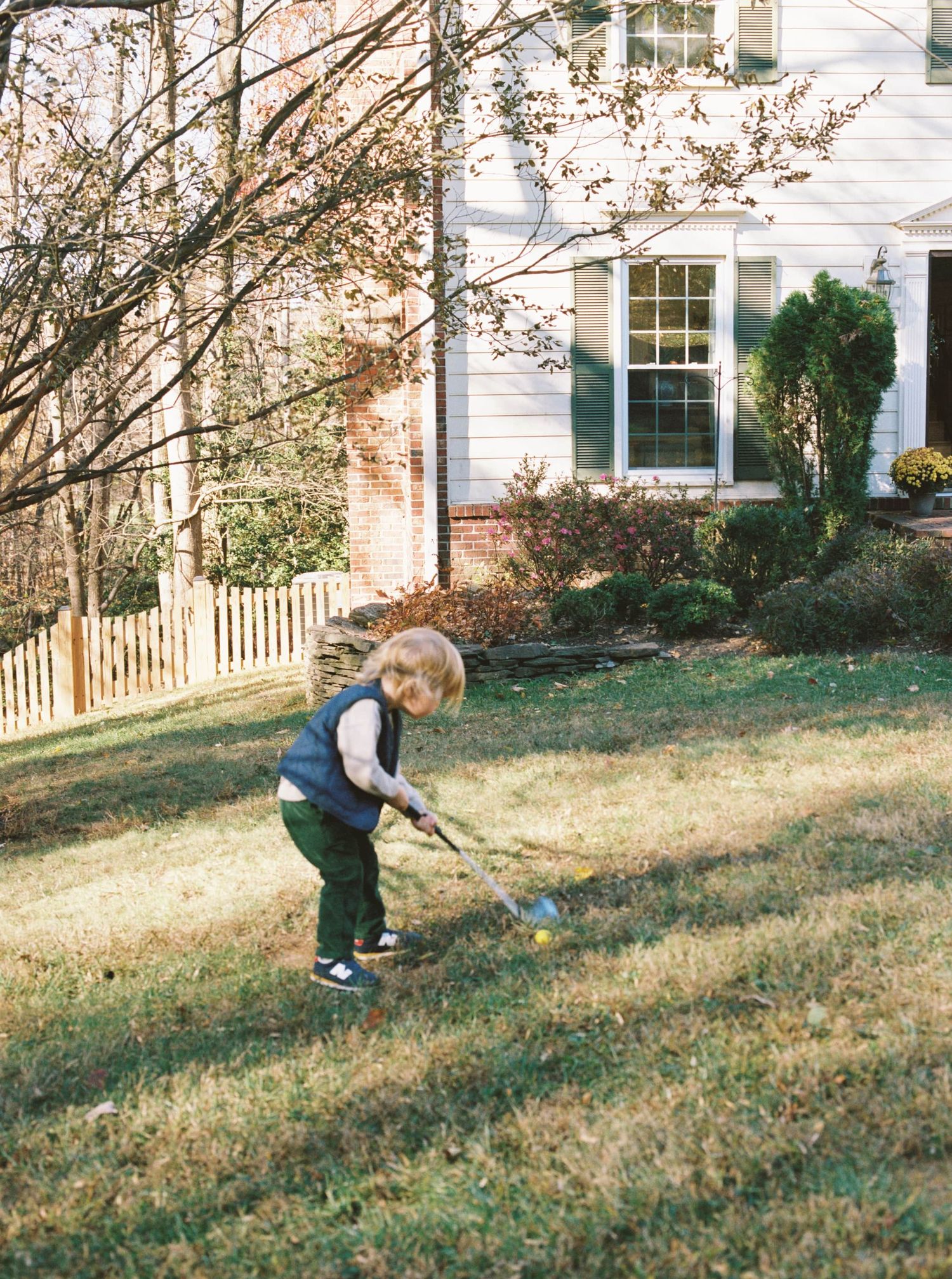 A small child plays in the yard of a white house with a picket fence on an autumn day.