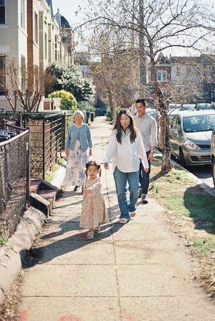 Asian family of four walking on a sunny sidewalk in a residential neighborhood, including grandparent, parents, and young child.