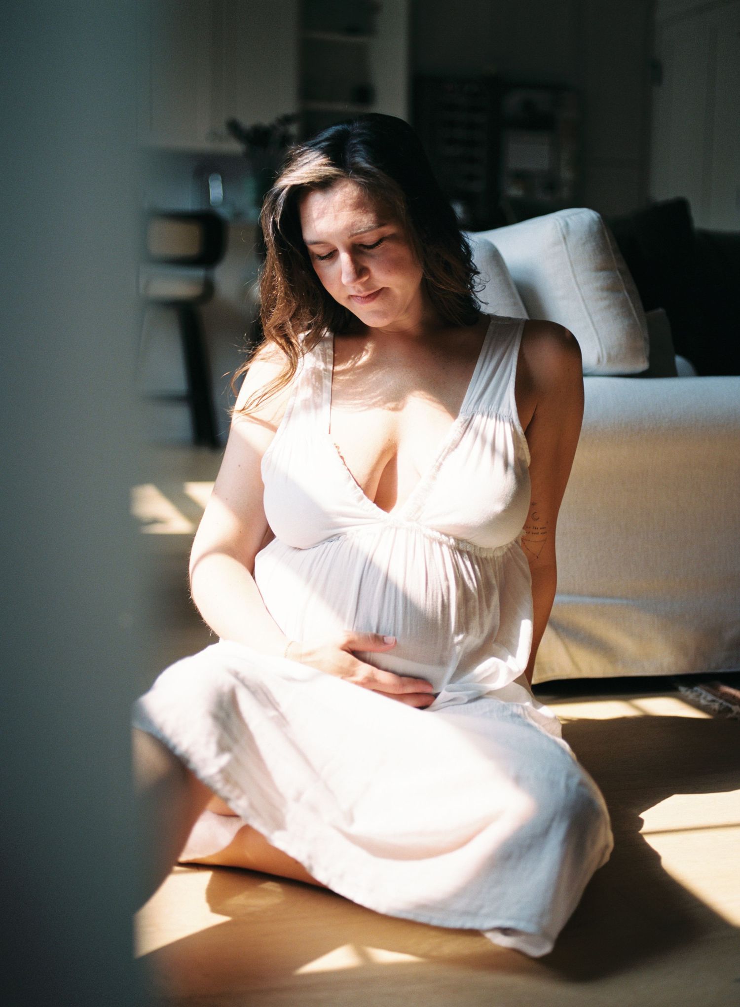 Pregnant woman in white dress sitting on floor in sunlight, cradling her baby bump with eyes closed.