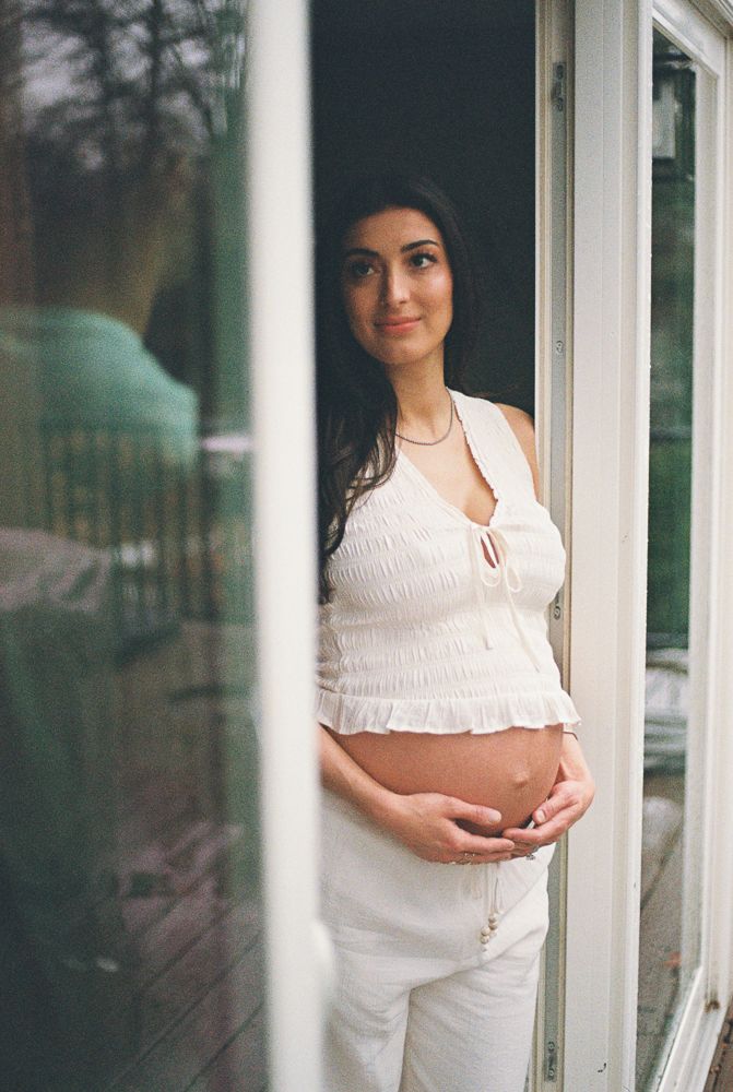 A pregnant woman in white clothing poses by a window during a maternity photoshoot.
