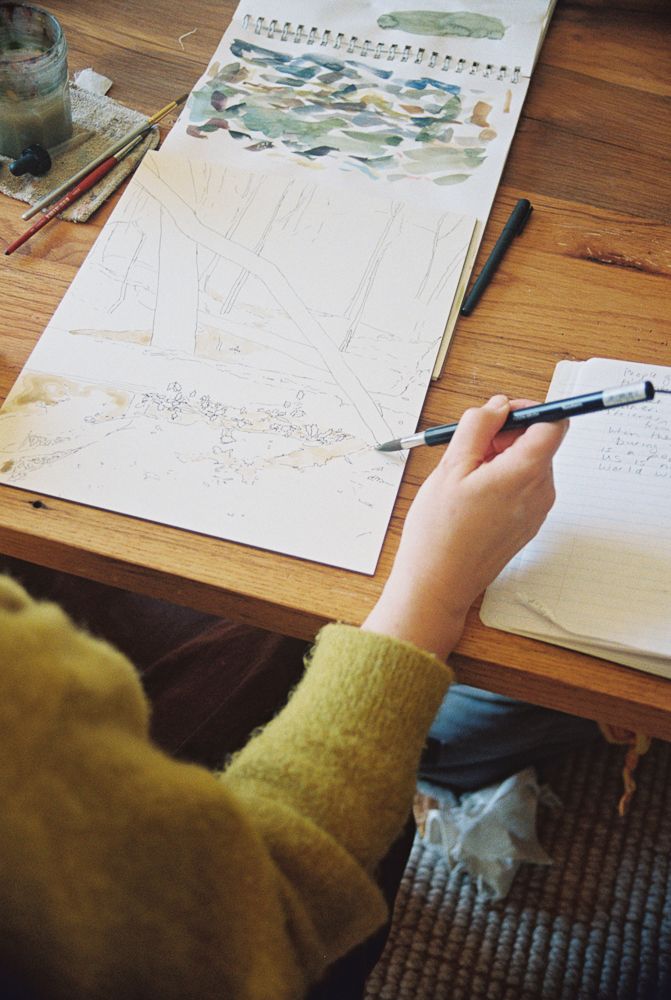 A hand in a yellow sweater writes in a notebook with watercolor artwork visible at the top of the page.