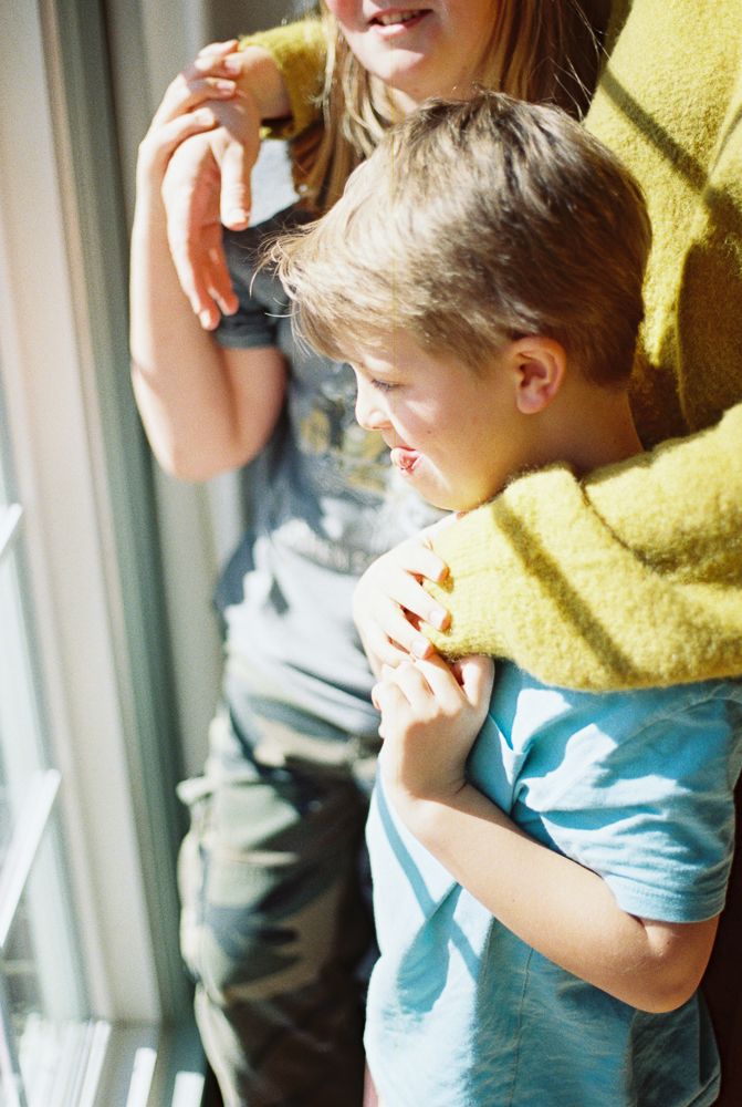 A child in a blue shirt and yellow sweater leans against a window while playing with a toy.