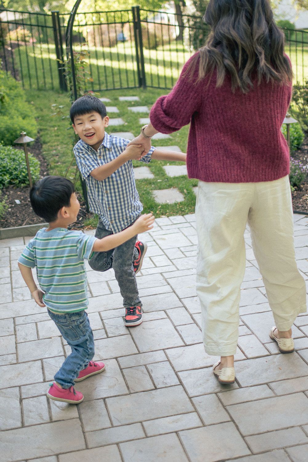 Two Asian boys playing with their mother in a garden patio, laughing and reaching out to hold hands.