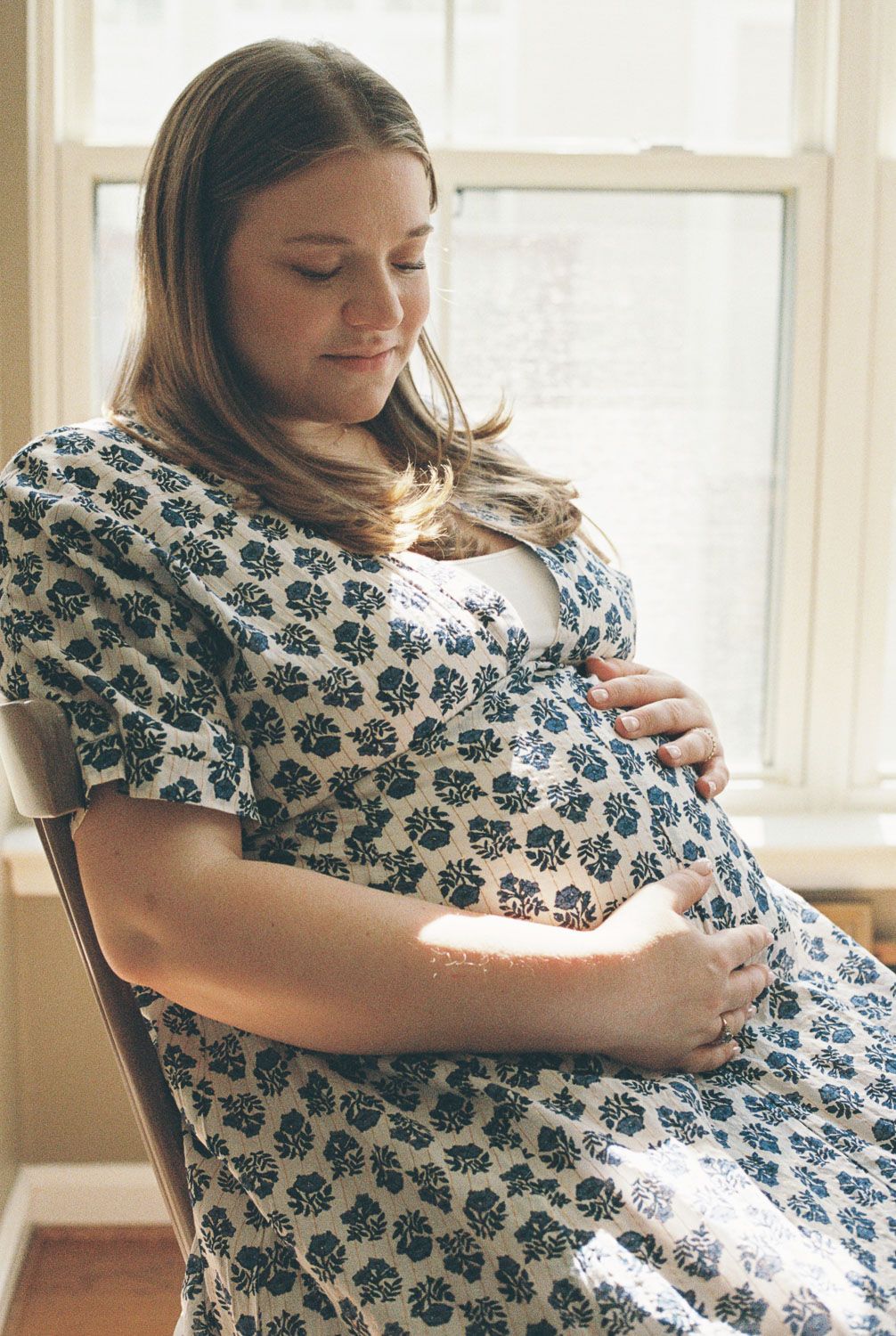 Pregnant woman in floral dress sitting in chair by window, cradling her baby bump with both hands.