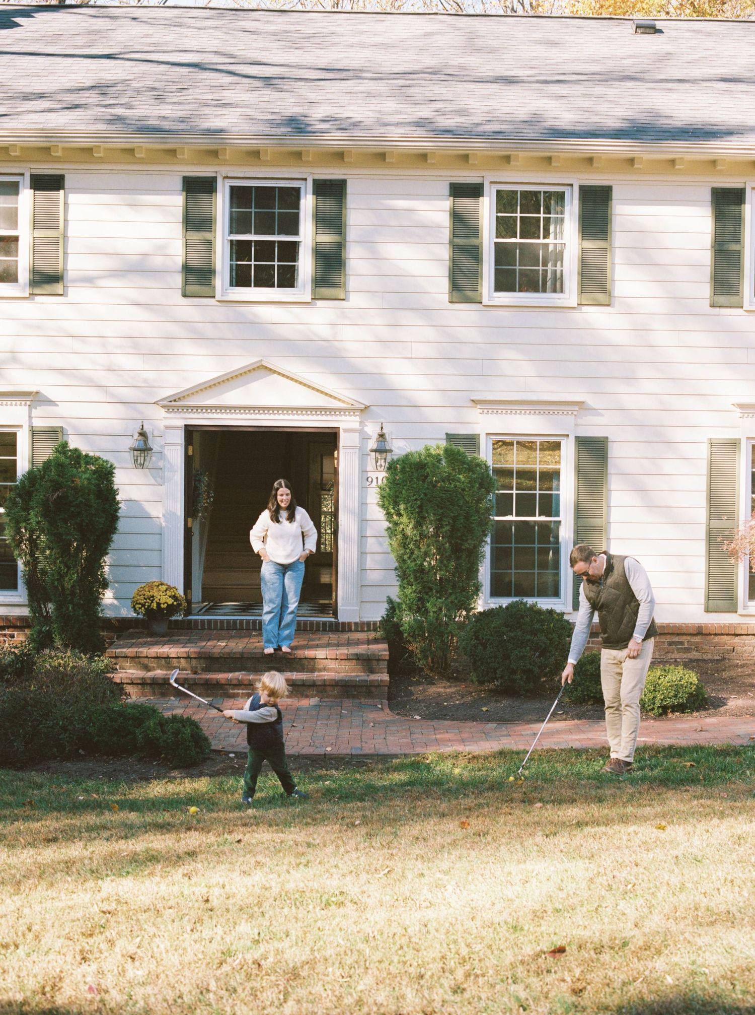 Three people in dark winter coats walk across a snow-covered lawn in front of a white colonial-style house.