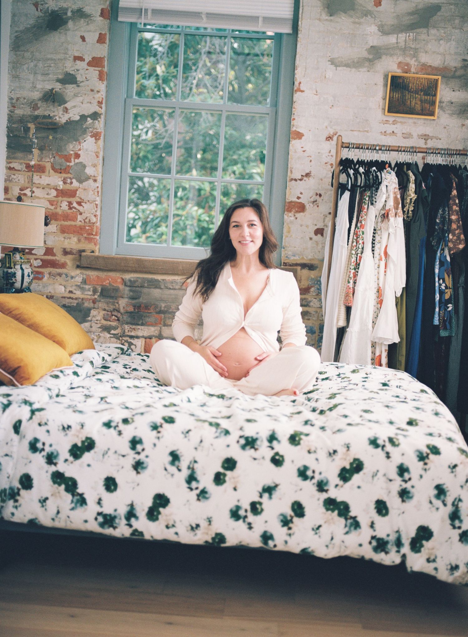 Someone relaxes on a white and floral print bedspread against a rustic exposed brick wall with windows.