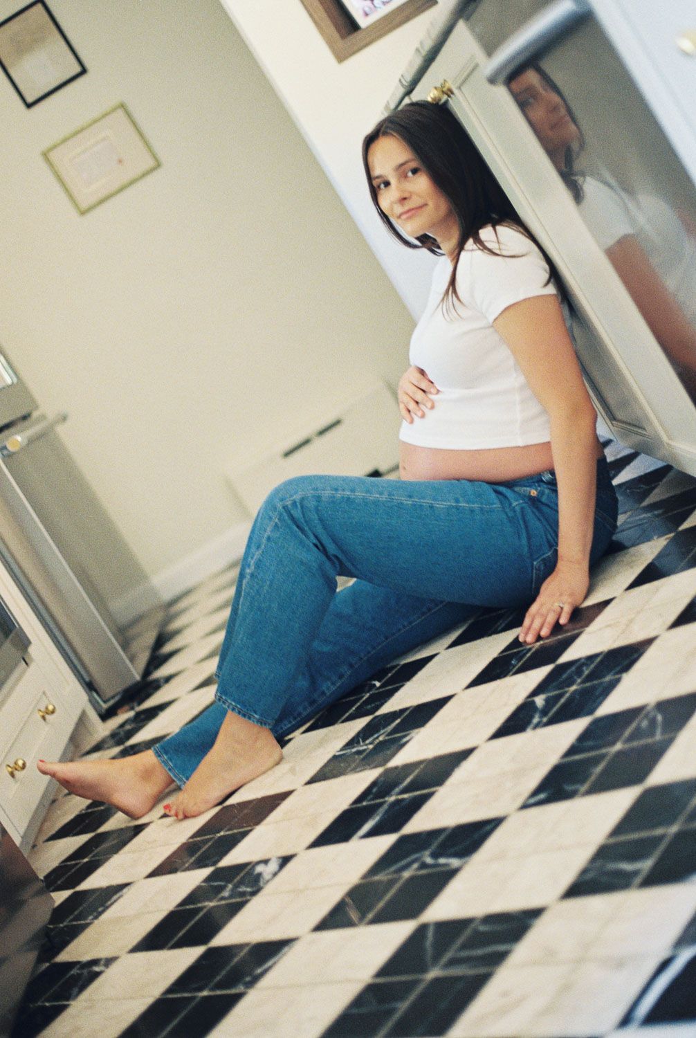 Pregnant woman in white crop top and jeans sitting barefoot on black and white checkered kitchen floor.