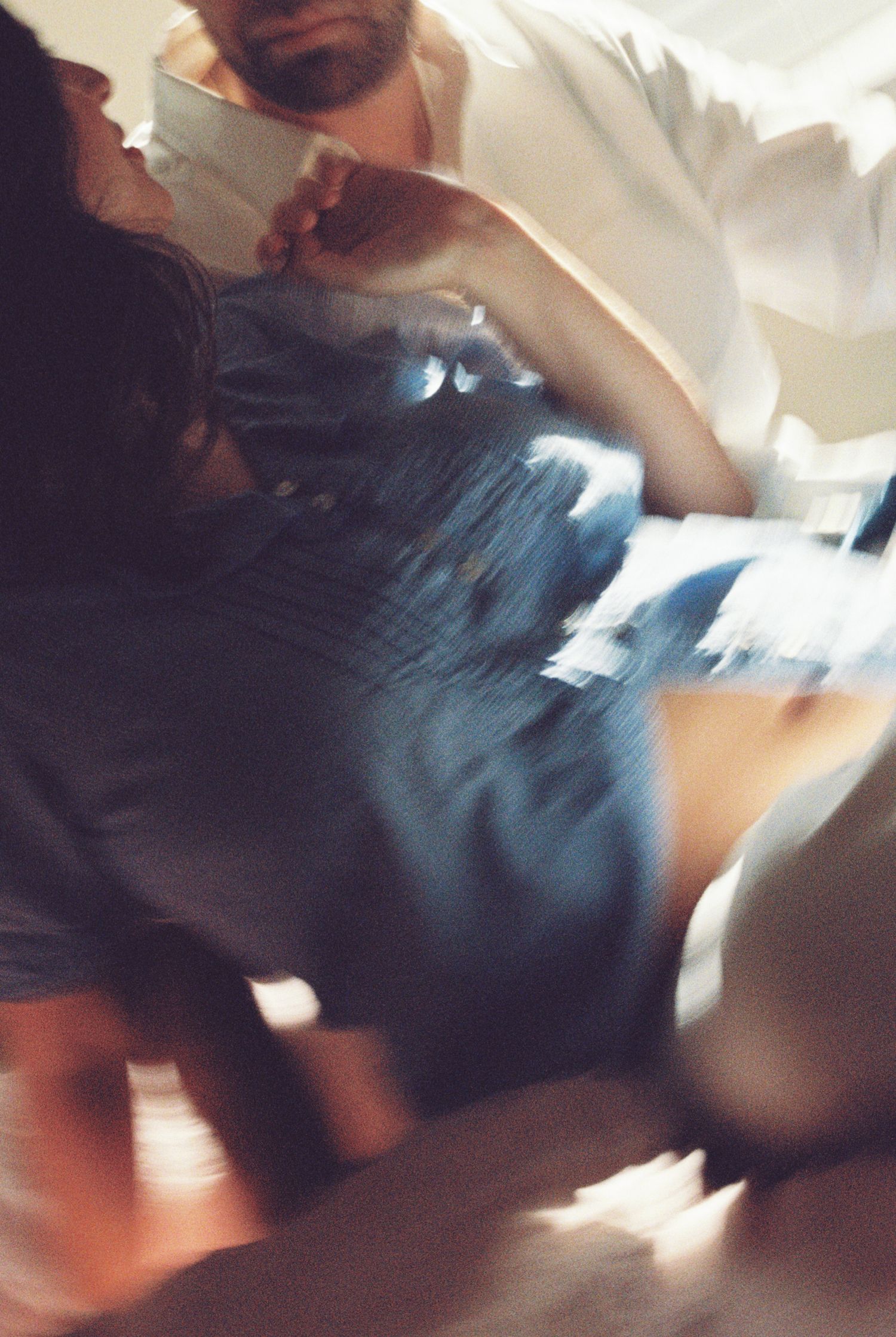Blurred, warm-toned photo of a bearded man in a white shirt leaning over a dark-haired woman in a denim dress in soft light.