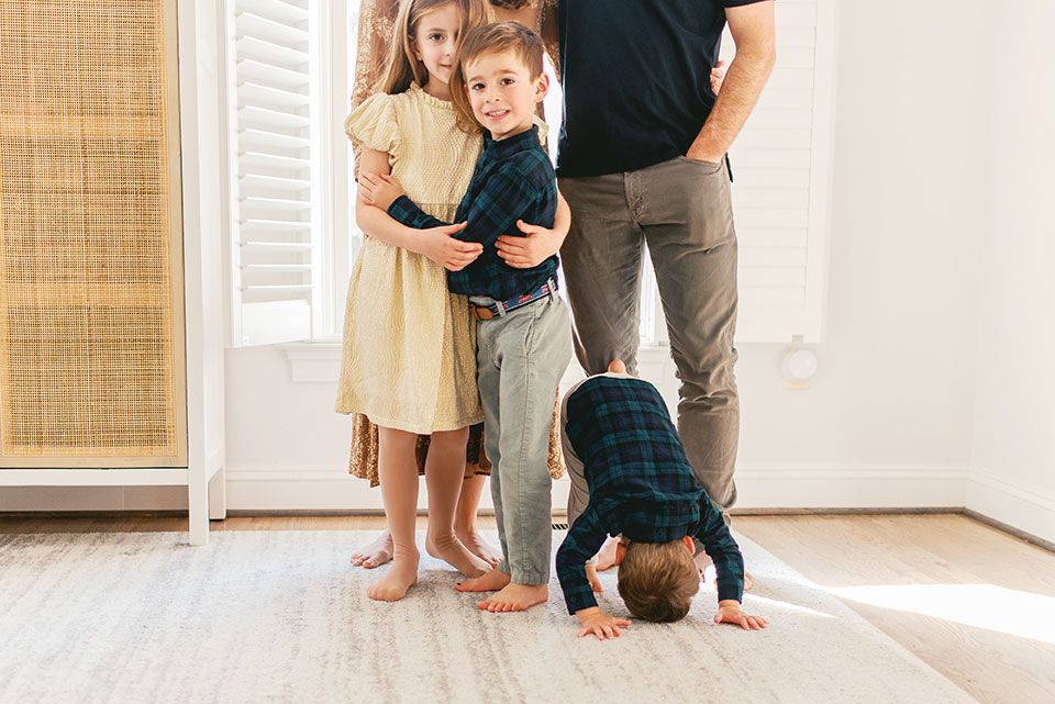 A family stands together in a bright room while a small child playfully bends over on a hardwood floor.