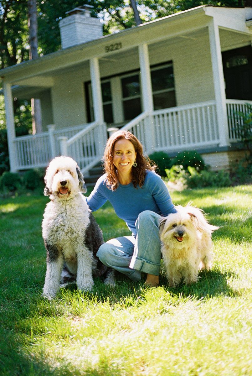 Woman kneeling on green lawn with two fluffy dogs in front of a white house with a covered porch.