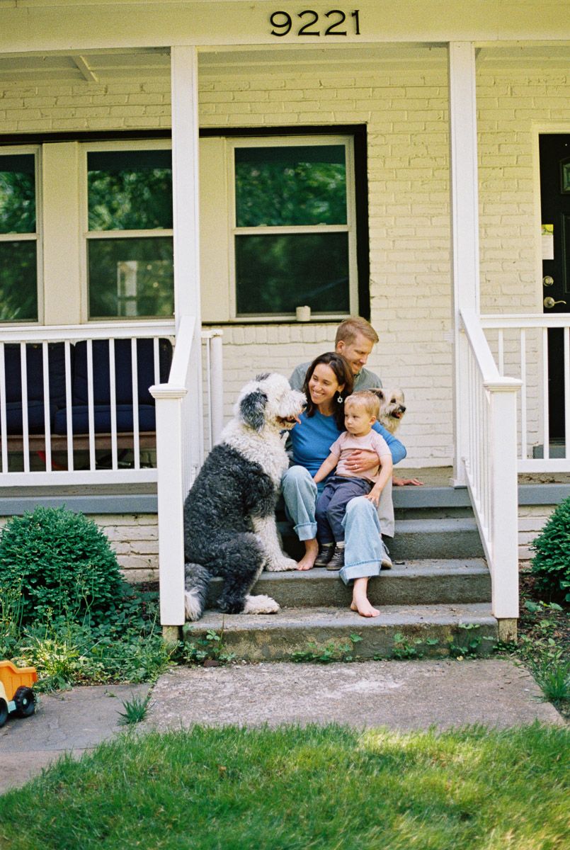 Family with toddler and two dogs sitting on front porch steps of white brick home numbered 9221.