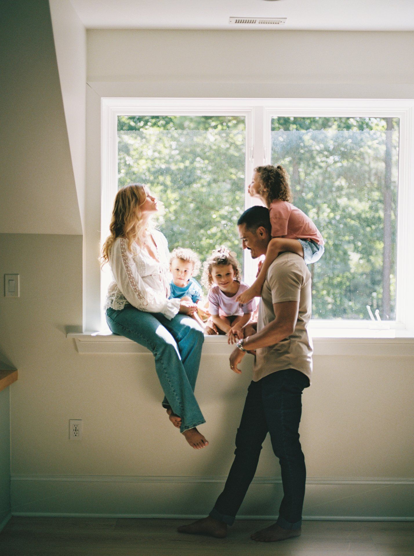 A family shares playful moments together in a bright sunlit room with white walls and windows.