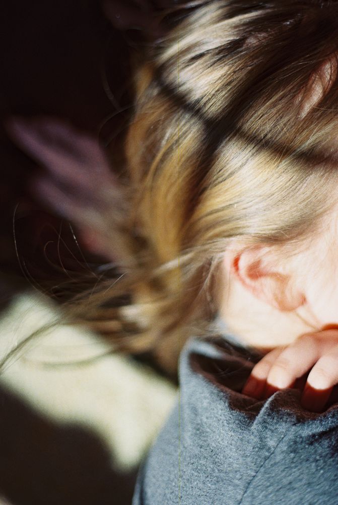 Close-up side view of blonde hair and ear with soft natural lighting and gray fabric in frame.