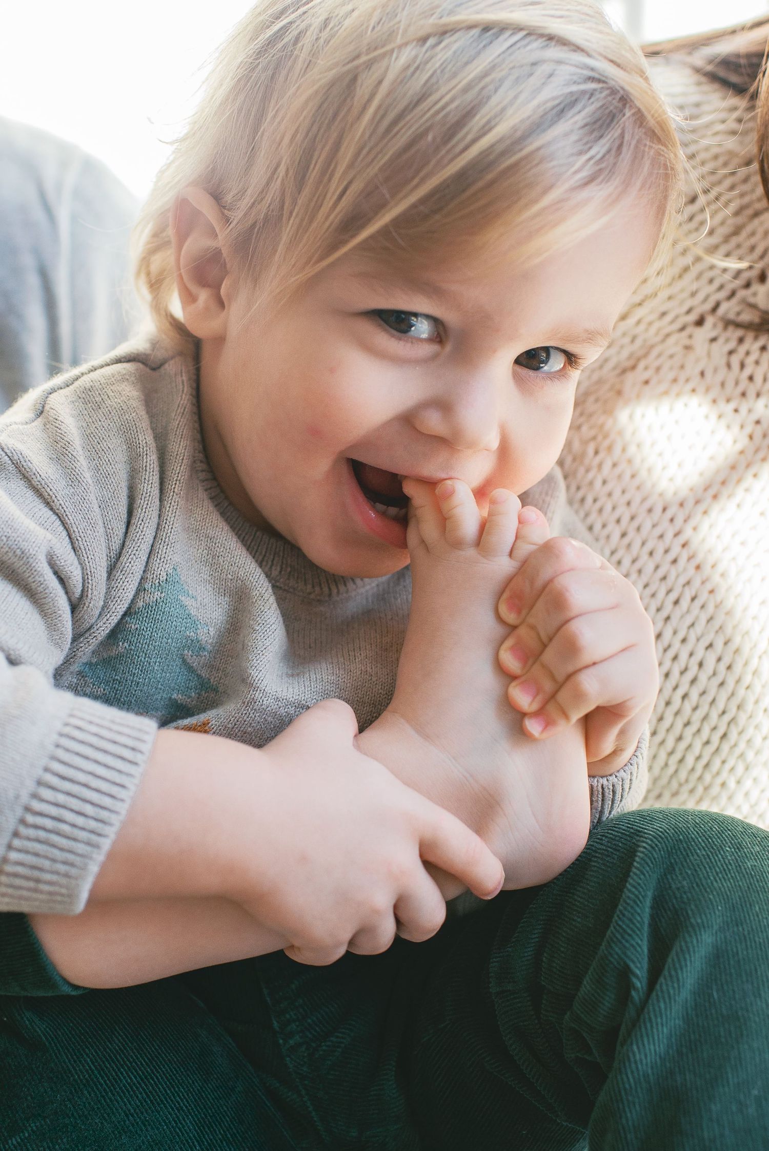 A young child in a gray sweater smiles joyfully while playfully biting their fingers.