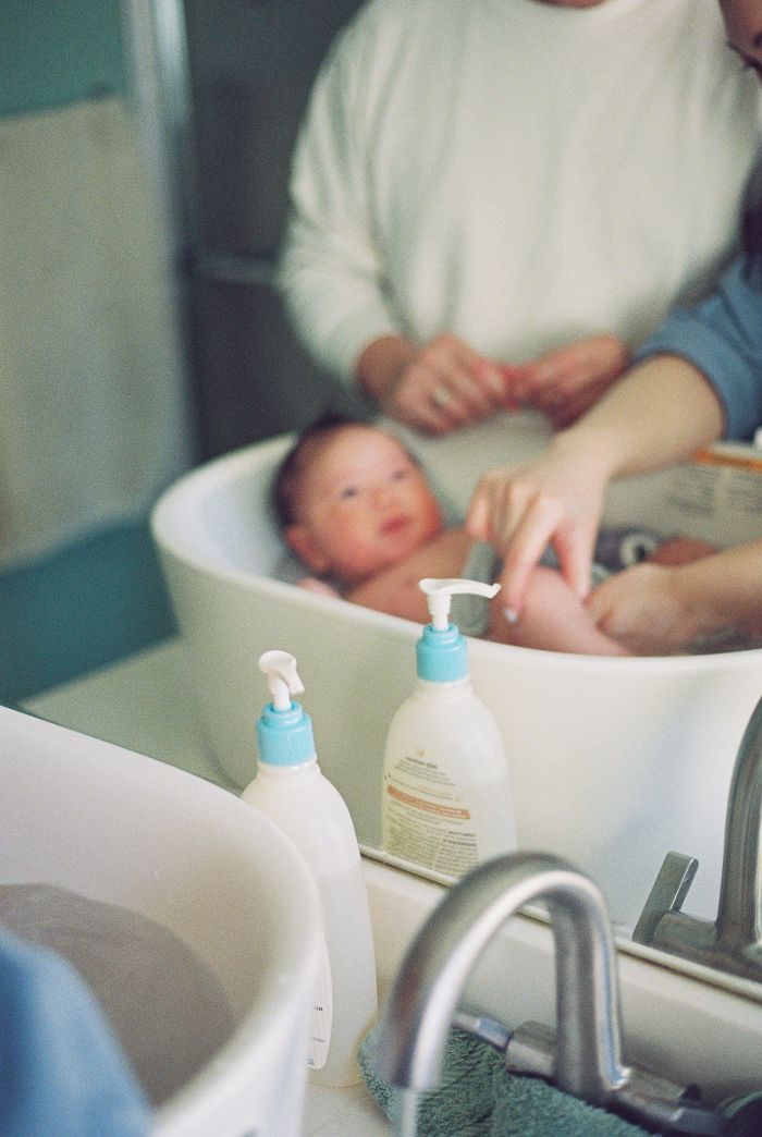 Parent bathing a newborn baby in a small white tub with baby wash bottles and a sink faucet in the foreground.