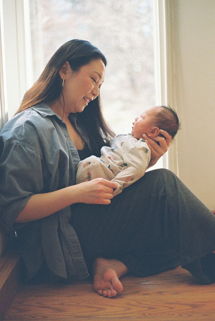Mother sitting by window smiling at her sleeping newborn baby.