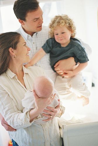A happy family with parents holding a newborn baby and a curly-haired toddler in a bright room.