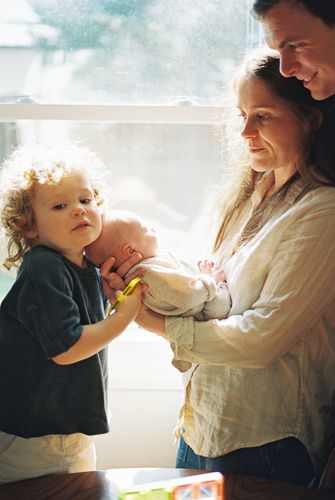 A young family with curly-haired toddler and parents lovingly holding a newborn baby by a bright window.