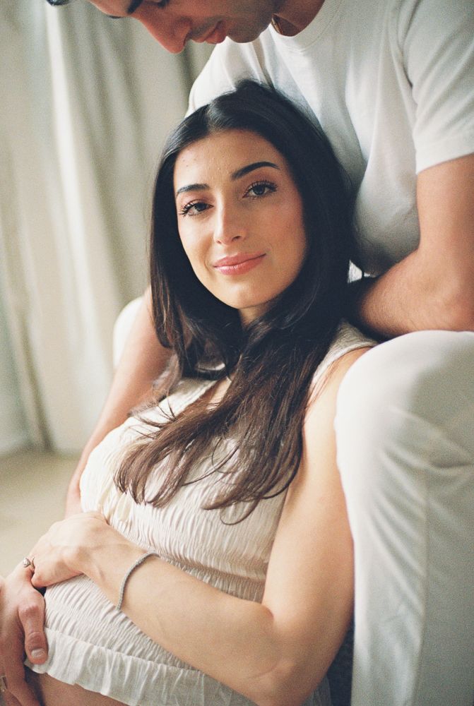 A woman in a white crop top relaxes against a light background while someone stands nearby.