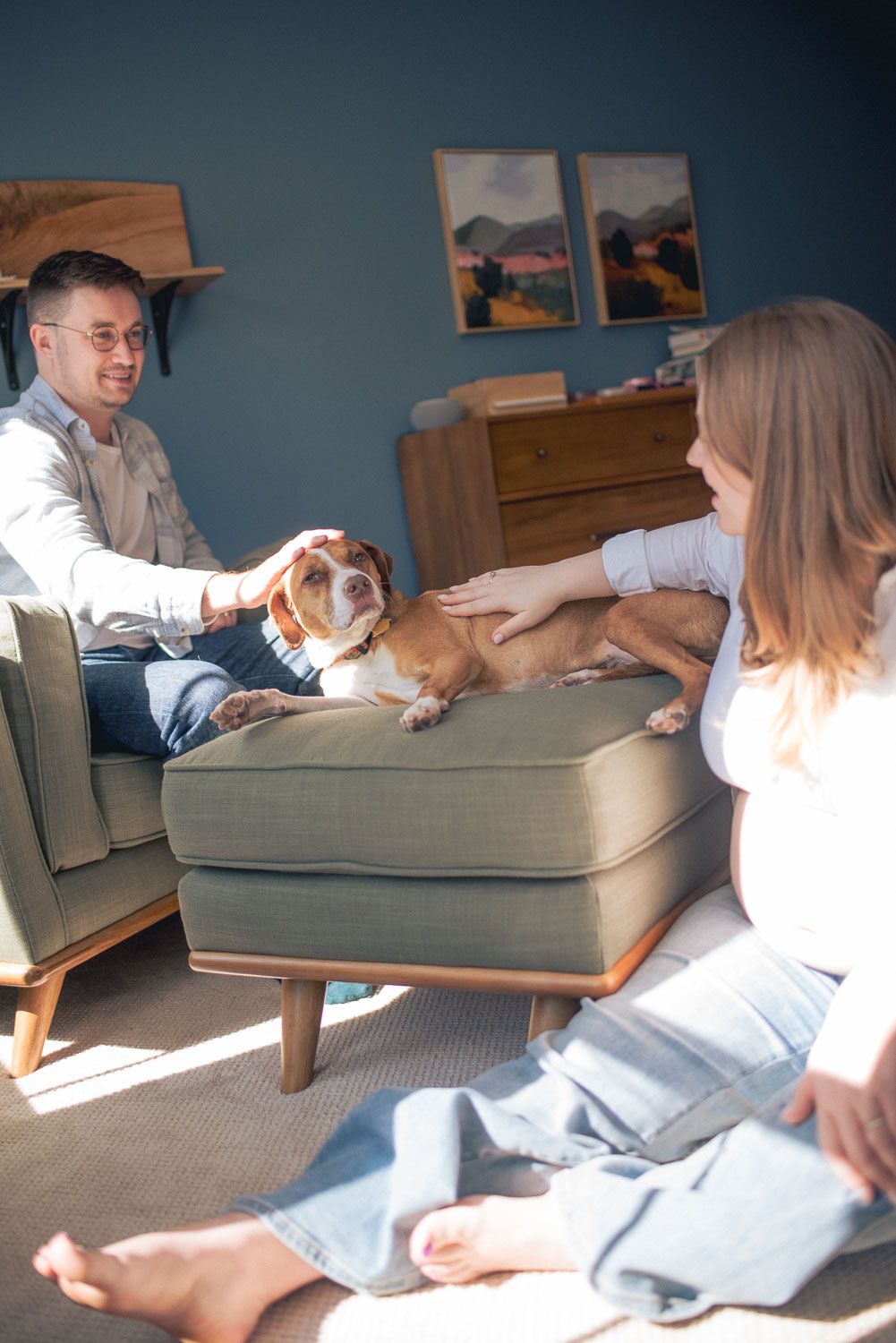 A couple pets their beagle dog resting on a green ottoman in a cozy, sunlit living room with blue walls.