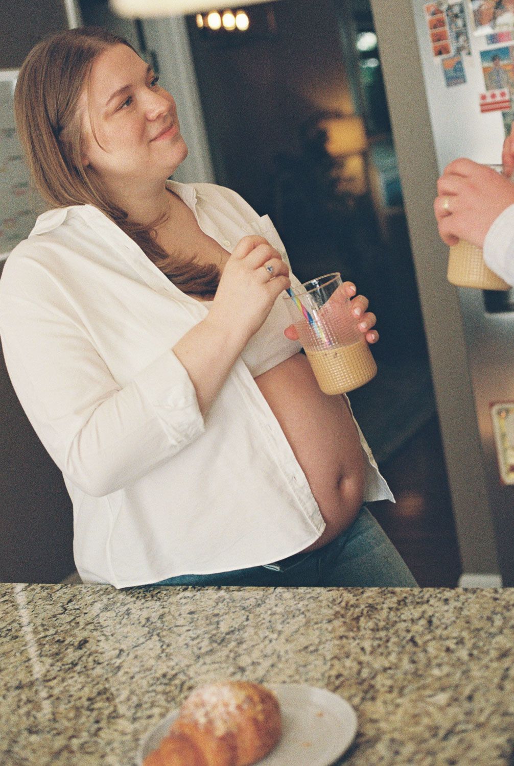 Pregnant woman in white shirt holding juice glass at kitchen counter with croissant on plate nearby.